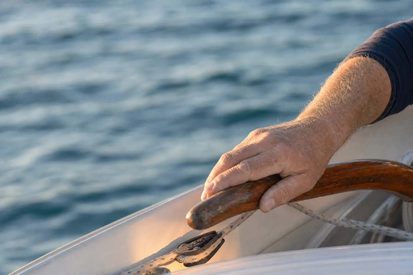 A weathered hand resting on a varnished timber tiller out on the water