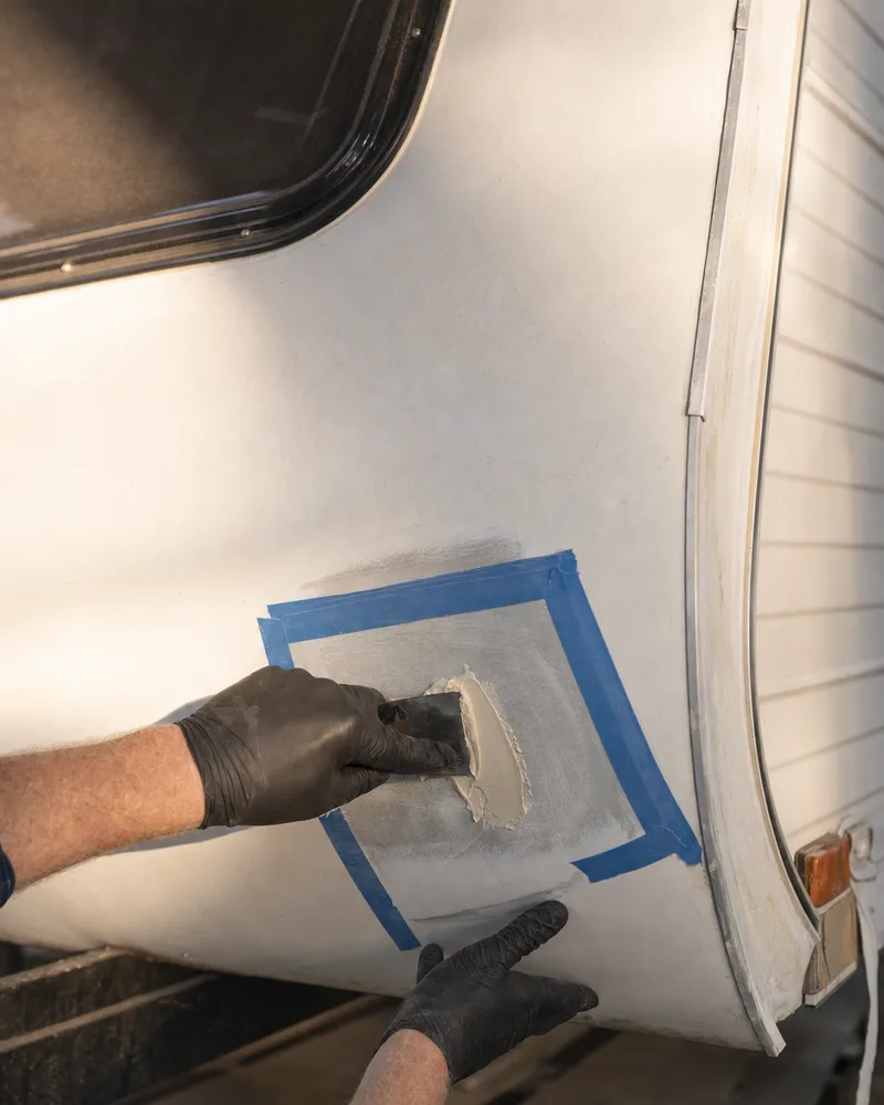 Gloved hands spreading filler over a repaired crack in the fibreglass corner of a caravan
